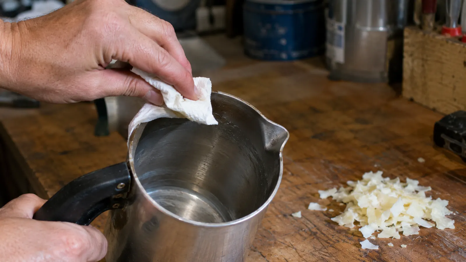 A candle maker wiping a metal pouring pot with a paper towel on a garage workbench